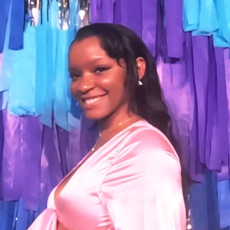 Rashida Darby, administrative operations and automation specialist, smiling in a pink satin top against a backdrop of layered blue and purple streamers.